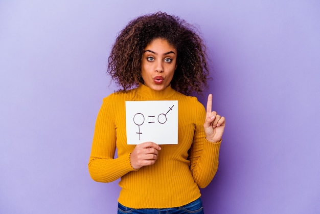 Young African American woman holding a gender equality placard isolated on purple wall having some great idea, concept of creativity.