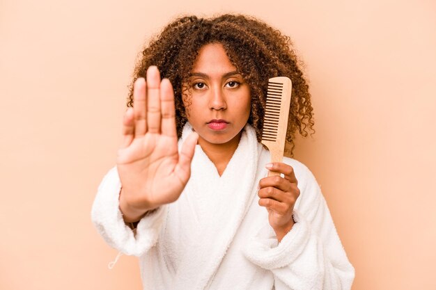 Young african american woman holding hairbrush isolated on\
beige background standing with outstretched hand showing stop sign\
preventing you