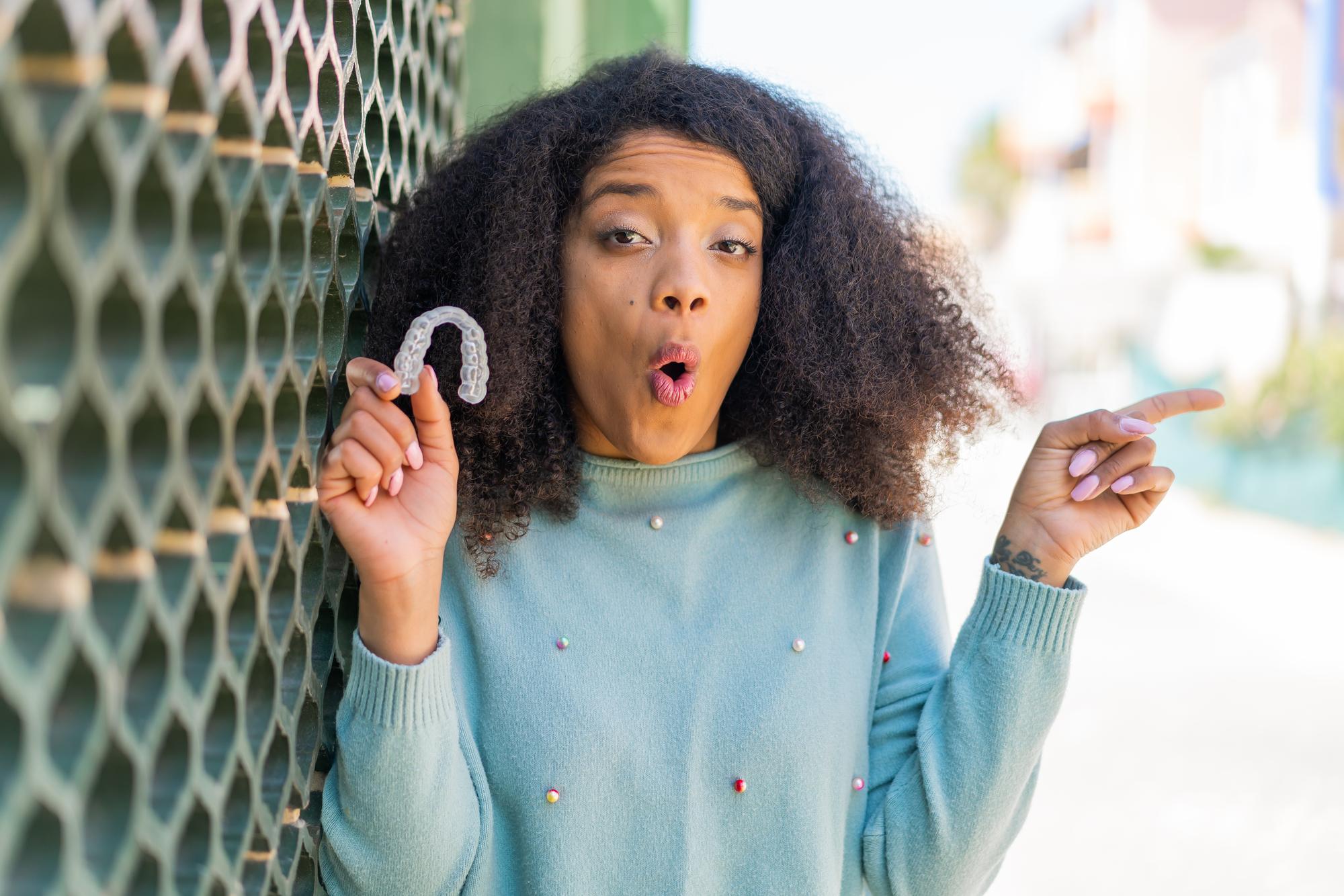 Foto giovane donna afroamericana che tiene un apparecchio dentale invisibile all'aperto sorpreso e puntando a lato