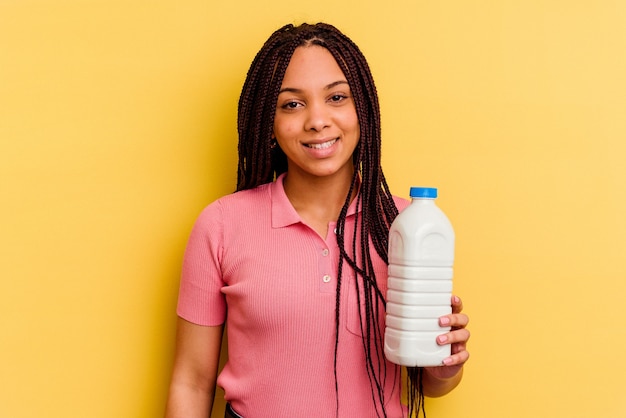 Молодая афро-американская женщина, держащая бутылку молока, изолированную на желтом фоне, счастливая, улыбающаяся и веселая.