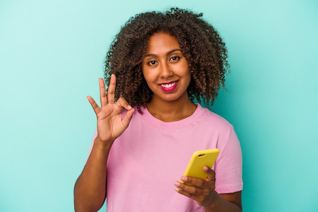 Giovane donna afroamericana che tiene un telefono cellulare isolato su fondo blu allegro e sicuro che mostra gesto giusto.