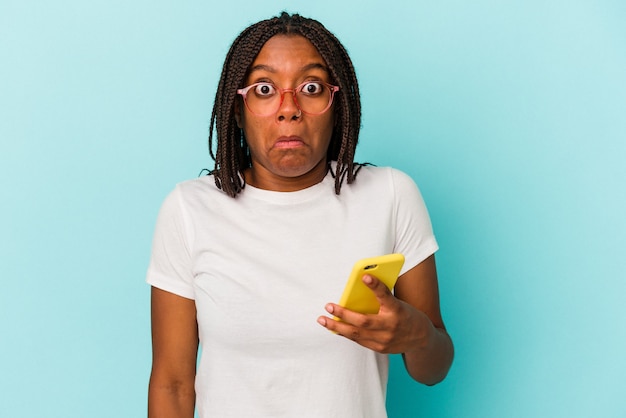 Young African American woman holding a mobile phone isolated on blue background  shrugs shoulders and open eyes confused.