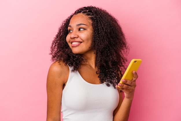 Young african american woman holding a mobile phone isolated a pink background looks aside smiling, cheerful and pleasant.