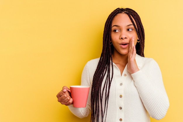 Young african american woman holding a mug isolated is saying a secret hot braking news and looking aside