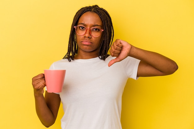嫌いなジェスチャーを示す黄色の背景に分離されたマグカップを保持している若いアフリカ系アメリカ人の女性は、親指を下に向けます。不一致の概念。
