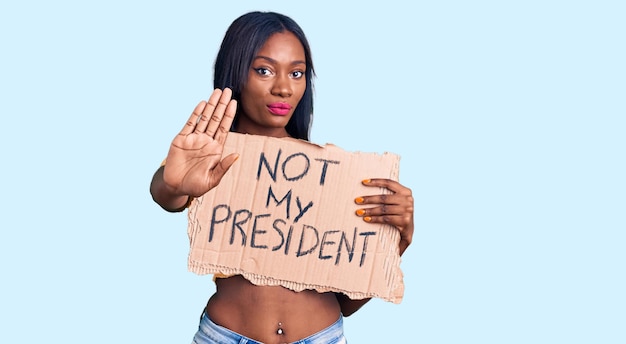 Young african american woman holding not my president protest banner with open hand doing stop sign with serious and confident expression, defense gesture