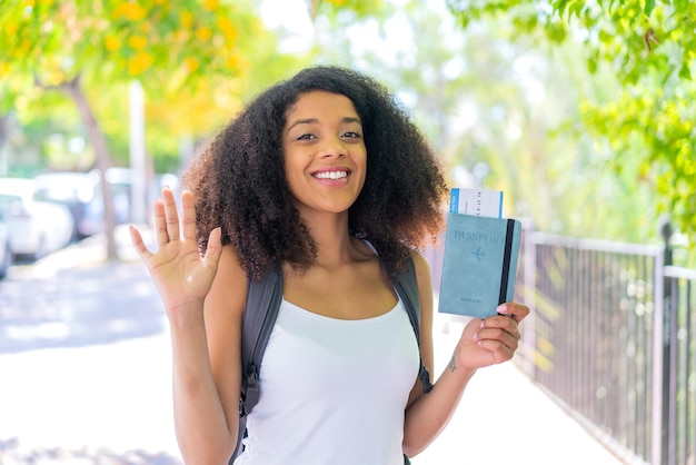 Young African American woman holding a passport at outdoors saluting with hand with happy expression