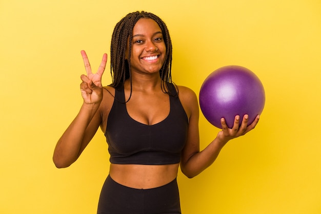 指で2番目を示す黄色の背景に分離されたピラティスボールを保持している若いアフリカ系アメリカ人女性。