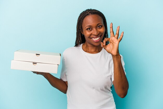 青の背景に分離されたピザを持っている若いアフリカ系アメリカ人女性は、明るく自信を持って大丈夫なジェスチャーを示しています。