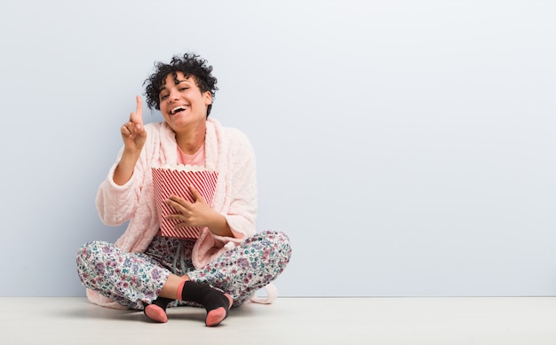 Young african american woman holding a popcorn bucket showing number one with finger.