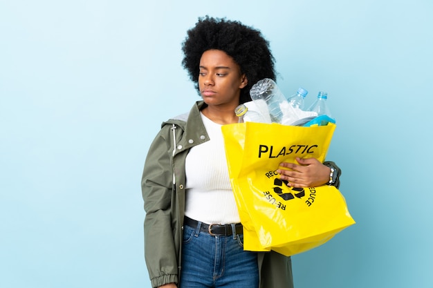 Young African American woman holding a recycle bag isolated on colorful having doubts while looking side