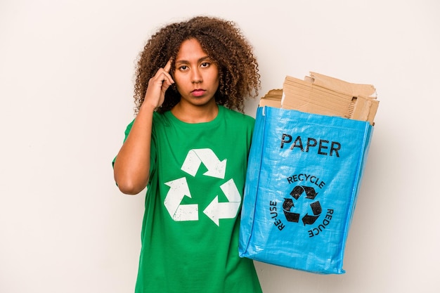 Молодая афроамериканка, держащая мешок для переработки, полный бумаги для переработки, изолирована на белом фоне, указывая пальцем на висок, думая, что сосредоточена на задаче