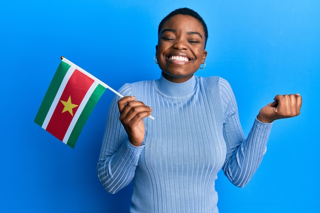 Young african american woman holding suriname flag screaming proud, celebrating victory and success very excited with raised arm