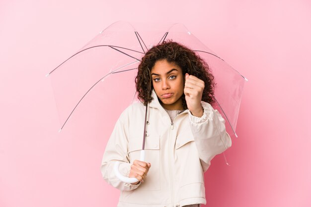 Young african american woman holding a umbrella showing fist to with aggressive facial expression.