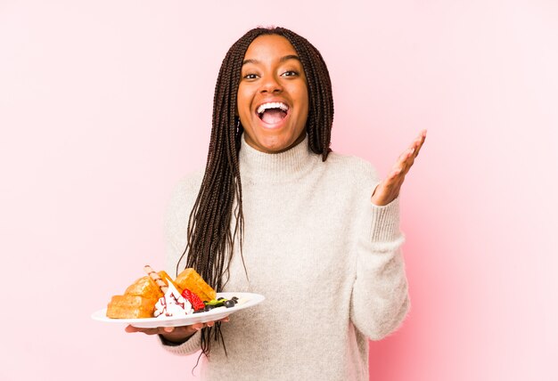 Young african american woman holding a waffle isolated receiving a pleasant surprise, excited and raising hands.