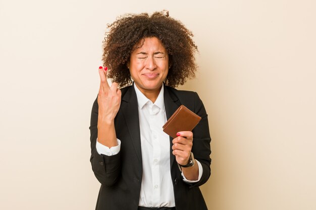 Young african american woman holding a wallet crossing fingers for having luck