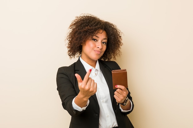 Young african american woman holding a wallet pointing with finger at you as if inviting come closer.