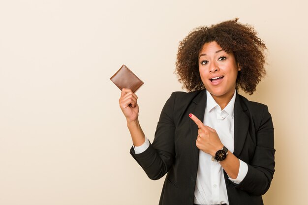 Young african american woman holding a wallet smiling cheerfully pointing with forefinger away.