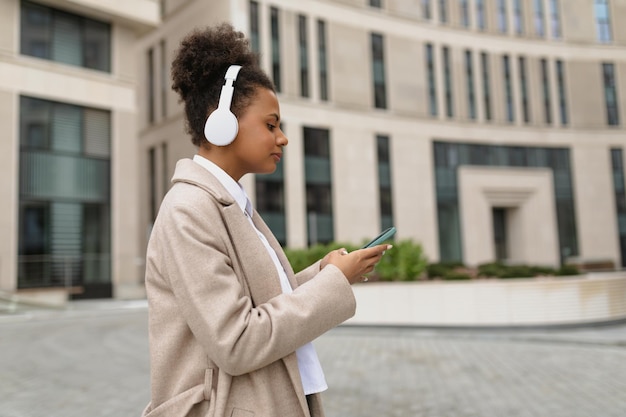 Photo young african american woman hurrying to work listening to music in headphones via mobile phone