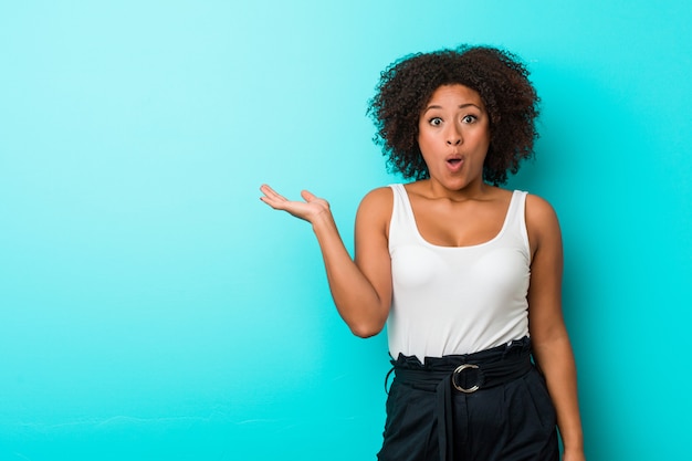 Young african american woman impressed holding copyspace palm.