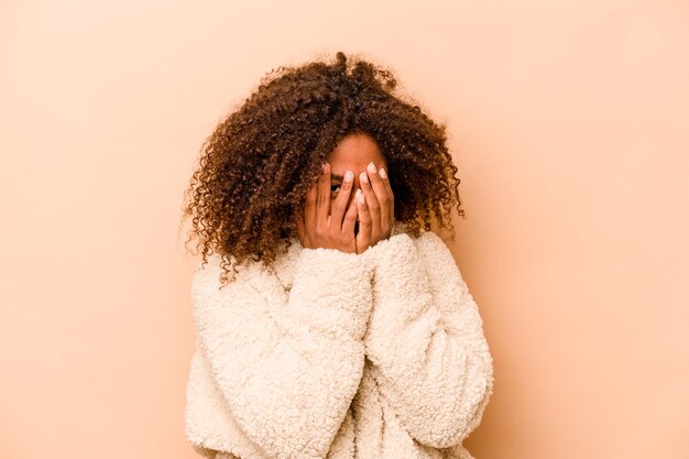 Young African American woman isolated on beige background blink at the camera through fingers embarrassed covering face