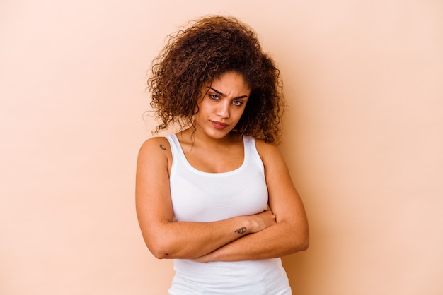 Young african american woman isolated on beige background frowning face in displeasure, keeps arms folded.