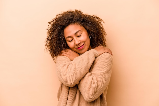 Young African American woman isolated on beige background hugs smiling carefree and happy