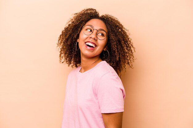 Young African American woman isolated on beige background looks aside smiling cheerful and pleasant