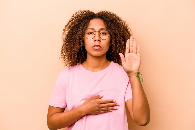 Young African American woman isolated on beige background taking an oath putting hand on chest