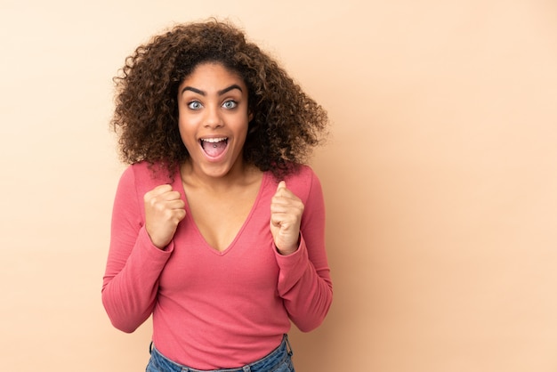 Giovane donna afroamericana isolata su spazio beige che celebra una vittoria nella posizione del vincitore