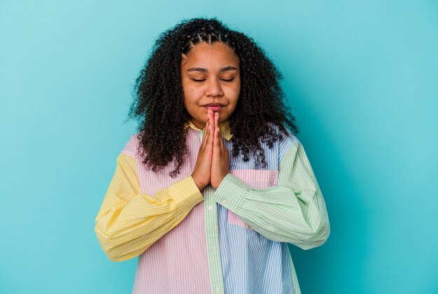 Young african american woman isolated on blue background holding hands in pray near mouth, feels confident.