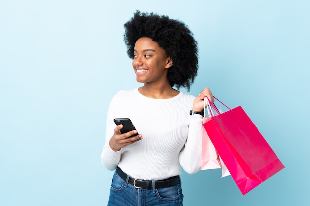 Young African American woman isolated on blue background holding shopping bags and a mobile phone