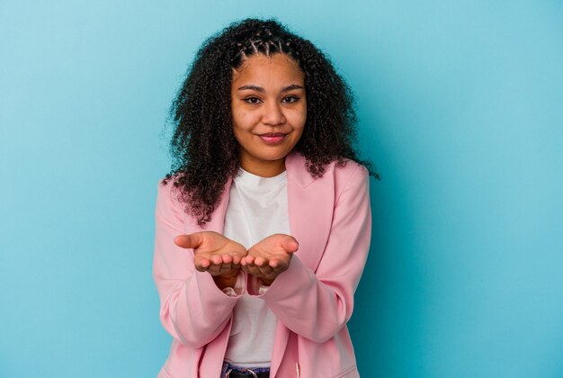 Young african american woman isolated on blue background holding something with palms, offering to camera.