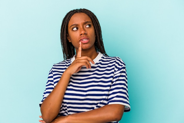Молодая афро-американская женщина, изолированная на синем фоне, смотрит в сторону с сомнительным и скептическим выражением лица.