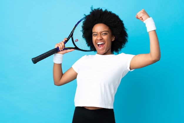 Giovane donna afro-americana isolata su sfondo blu, giocare a tennis e celebrare una vittoria