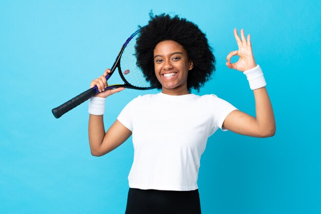 Giovane donna afroamericana isolata su fondo blu che gioca a tennis e che fa segno giusto