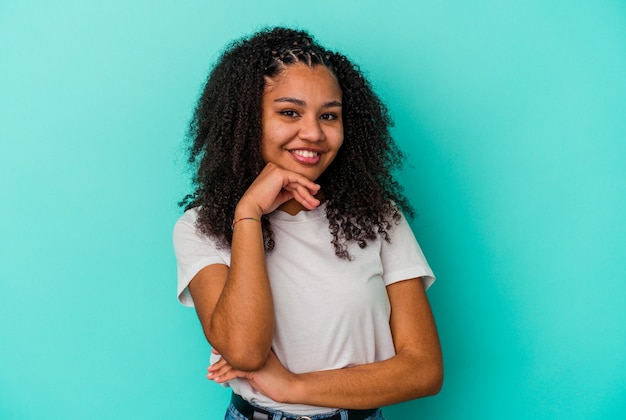 Молодая афро-американская женщина, изолированных на синем фоне, улыбается, счастлива и уверенно, трогательно подбородок рукой.