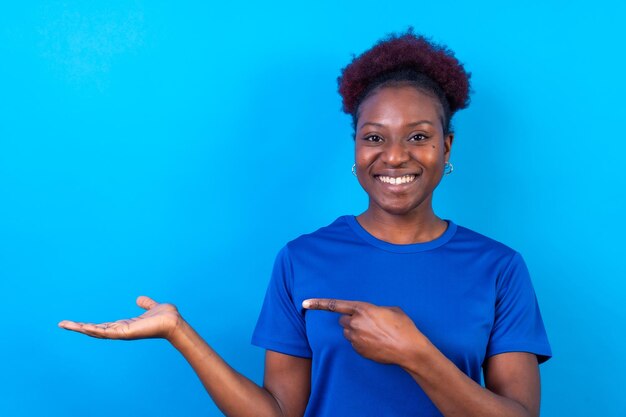 Young african american woman isolated over blue background smiling and pointing studio shoot copy space