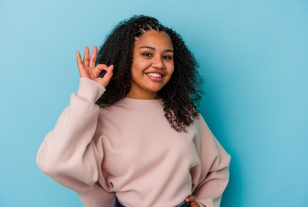 Young african american woman isolated on blue background winks an eye and holds an okay gesture with hand.