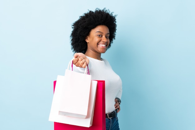 Photo young african american woman isolated on blue holding shopping bags and giving them to someone