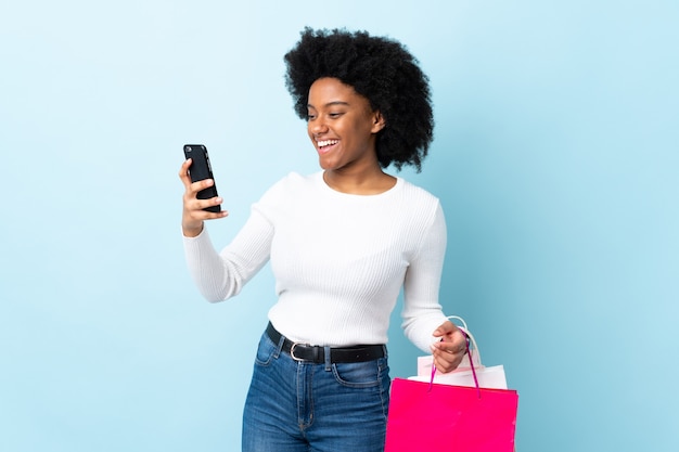 Young African American woman isolated on blue holding shopping bags and writing a message with her cell phone to a friend