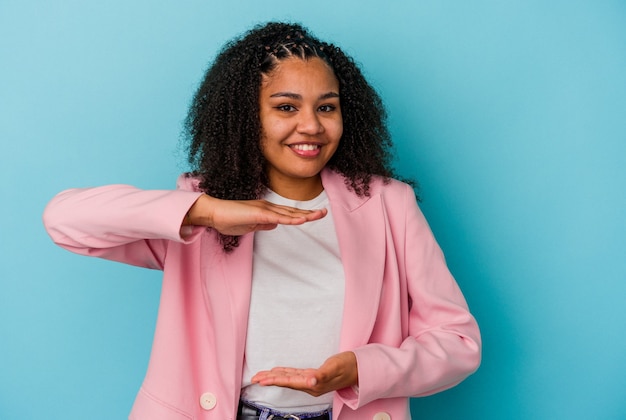 Young african american woman isolated on blue wall holding something with both hands, product presentation