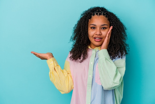 Young african american woman isolated on blue wall holds copy space on a palm, keep hand over cheek. Amazed and delighted.