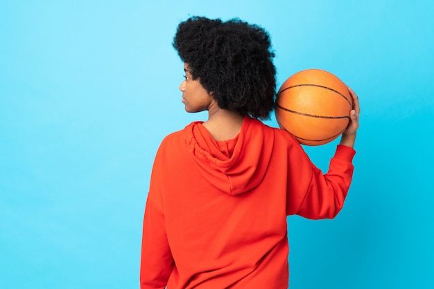 Young african american woman isolated on blue wall playing\
basketball in back position