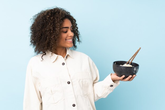 Young African American woman isolated on blue with happy expression while holding a bowl of noodles with chopsticks