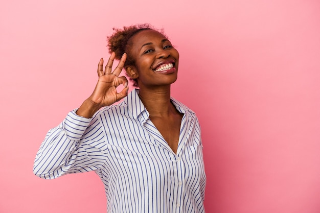 Young african american woman isolated on pink background winks an eye and holds an okay gesture with hand.