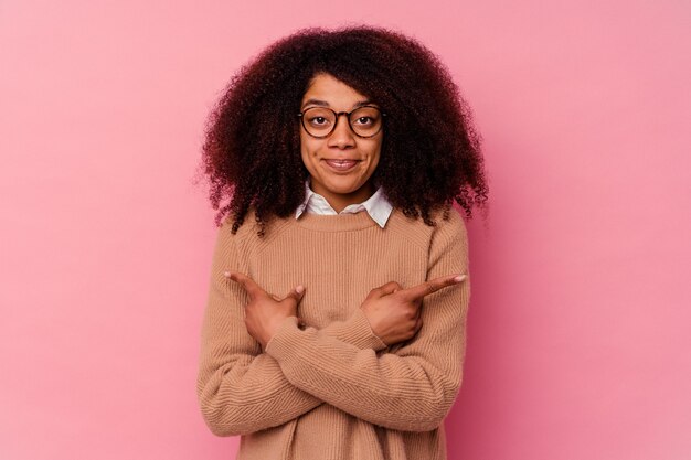 Young african american woman isolated on pink points sideways, is trying to choose between two options