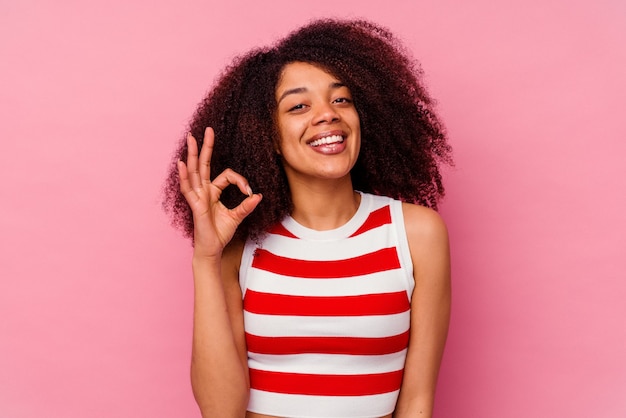 ピンクの壁に孤立した若いアフリカ系アメリカ人の女性は、陽気で自信を持って大丈夫なジェスチャーを示しています。