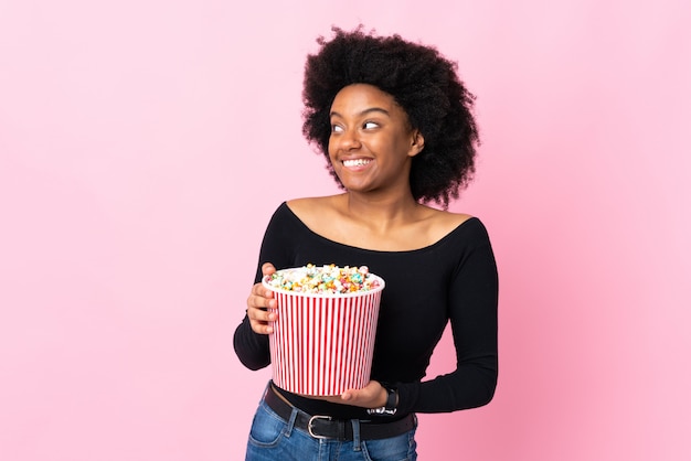 Young African American woman isolated on pink wall holding a big bucket of popcorns