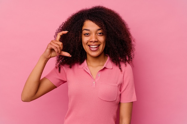 Young african american woman isolated on pink wall holding something little with forefingers, smiling and confident.